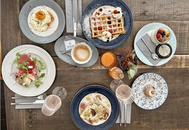 Ein schön gedeckter Tisch mit verschiedenen Gerichten zum Frühstück. Es sind Waffeln, Eier, Aufschnitt und Getränke wie Kaffee und Saft zu sehen.