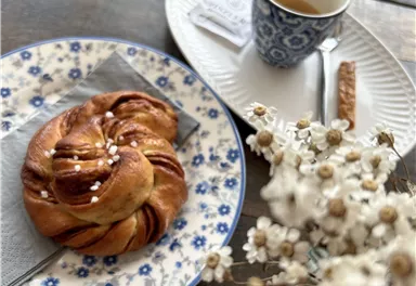 Ein süßer Hefeknoten auf einem blauen Teller mit einer Tasse Kaffee daneben. Im Hintergrund steht eine kleine Vase mit zarten Blumen.