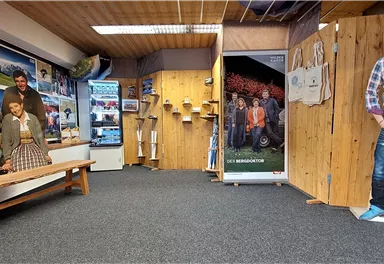 Ein Innenraum mit Holzwänden und mehreren Bildern an der Wand. Auf einer Bank sitzen geschnittene Figuren von Personen, und eine weitere Figur steht daneben.