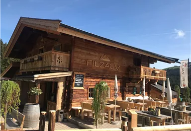 A cozy wooden house with an outdoor restaurant. There is a terrace with tables and sun umbrellas in a mountain landscape.