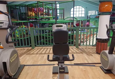A fitness room with several training machines and a playful area in the background. The room is brightly lit and inviting.