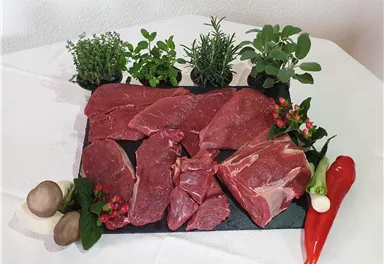 Fresh meat on a black stone board, surrounded by various herbs like thyme and rosemary. Red and green bell peppers as well as mushrooms are arranged alongside.
