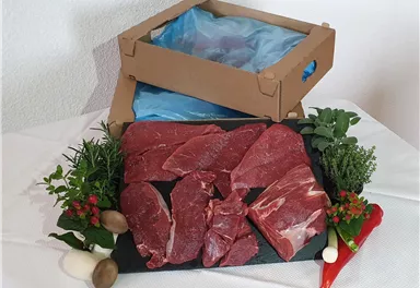 Fresh meat on a black granite board, surrounded by herbs and vegetables. In the background, an opened package stands.
