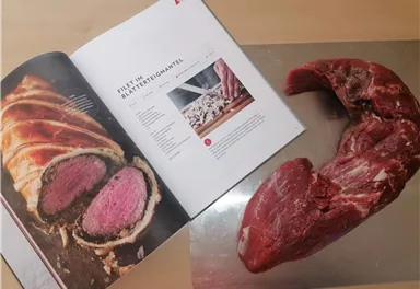 A fresh piece of meat lies next to a cookbook. The page displays a recipe for a fillet dish.