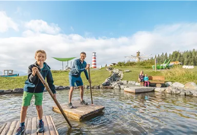 Zwei Kinder stehen auf einer Holzplattform im Wasser und haben Spaß. Im Hintergrund sind grüne Wiesen und ein Leuchtturm zu sehen.