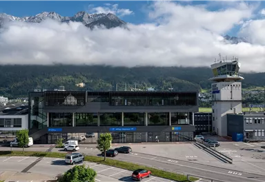 A modern airport with a control tower and surrounding mountains. The sky is partly cloudy, and the landscape is green.