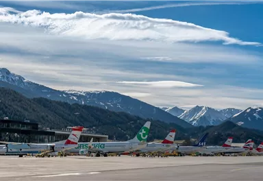 An airport with several airplanes on the tarmac and majestic mountains in the background. The sky is clear with a few clouds.