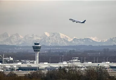 An airport with a control tower and a departing airplane. Snow-covered mountains are visible in the background.