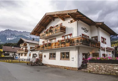 A charming two-story house with a wooden balcony and flowers. In the background, mountains and a quiet road can be seen.
