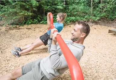 Two people are training outdoors on a fitness machine. They are pulling on a red bar on a surface covered with wood chips.