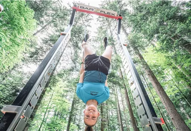 A young man is hanging upside down on a training device in the woods. The surroundings are green and filled with light.
