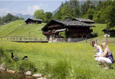 An idyllic country house with a double facade in a green meadow. A girl and a woman are watching ducks next to a small body of water.