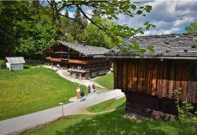 Two traditional wooden buildings in a green environment. A path leads through the landscape, and people are on the go.
