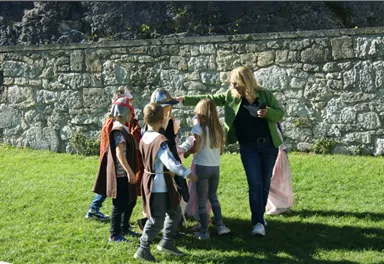Eine Gruppe von Kindern in Kostümen steht in einem Garten. Eine Erwachsene interagiert freundlich mit den Kindern.