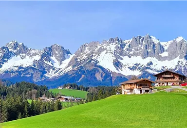 Eine malerische Berglandschaft mit schneebedeckten Gipfeln und saftig grünen Wiesen. Im Vordergrund sind traditionelle Holzhäuser und ein Weg zu sehen.