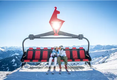 Two skiers are sitting on a chairlift in the mountains. The surroundings are covered in snow and the sun is shining clearly in the sky.