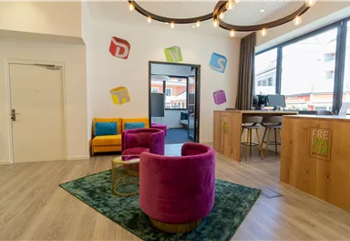 Ein modernes Empfangsbereich mit bunten Möbeln und einem freundlichen Ambiente. Die Sitzecke besteht aus lila Sesseln und einer Holztheke.