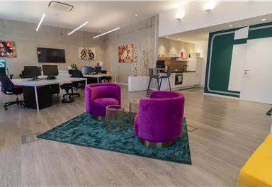 Ein modernes Büro mit bunten Sesseln und einem grünen Teppich. Es gibt mehrere Arbeitsplatzstationen und eine offene Küche im Hintergrund.