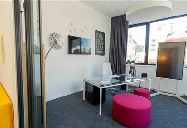 Ein modernes Büro mit einem transparenten Tisch und bunten Sitzmöglichkeiten. Große Fenster sorgen für viel Tageslicht.