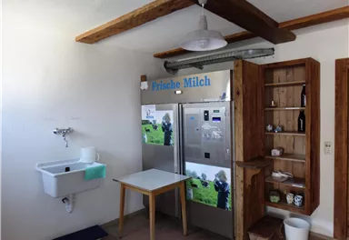 A cozy kitchen with a refrigerator offering fresh milk. A table and a wooden shelf complement the space.