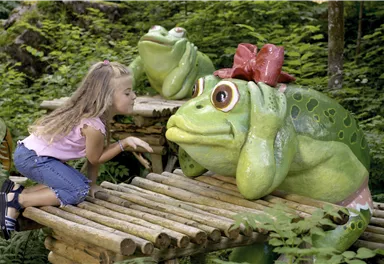 A girl kisses a large green plastic frog with a red bow. In the background, more frogs and a lot of green foliage can be seen.