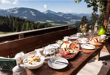 A beautiful outdoor table is set with a variety of dishes and drinks. In the background, majestic mountains and a green landscape are visible.