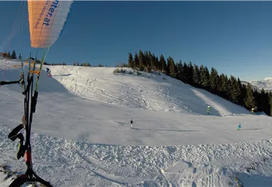 A snow-covered mountain landscape with a clear sky. In the foreground, a paraglider with a colorful parachute can be seen.