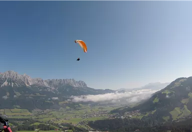 A paraglider hovers over a picturesque mountain landscape. The clear weather and the green meadows create a beautiful backdrop.
