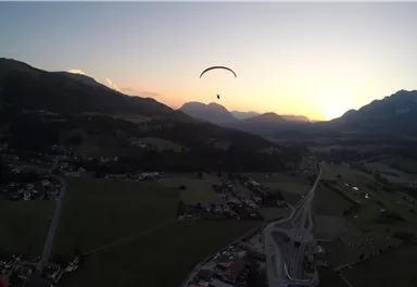 A paraglider floats over a landscape with mountains and a beautiful sunset in the background. The surroundings are tranquil with green meadows and small villages.