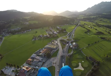 A breathtaking view from above of a green landscape with a small village and mountainous backdrop. The sun sets on the horizon, casting warm light over the scene.