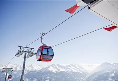A red gondola on a cable car glides over snow-covered mountains. The sky is clear and blue.