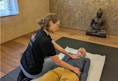 A woman is giving a treatment to a lying person in a quiet room. In the background, there is a Buddha statue that contributes to the relaxed atmosphere.