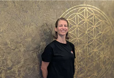 A smiling woman stands in front of a golden wall with a geometric pattern. She is wearing a black T-shirt and appears friendly and inviting.
