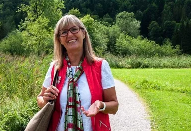 Eine freundliche Frau lächelt auf einem Wanderweg umgeben von grüner Natur. Im Hintergrund sind Bäume und eine sonnige Landschaft zu sehen.