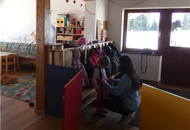 Ein heller Raum mit Holzmöbeln und bunten Spielmatten. Eine Erzieherin hilft einem Kind, sich anzuziehen.