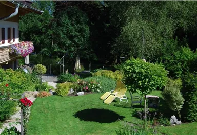 A beautiful garden with lush greenery and colorful flowers. Two lounge chairs are placed on the lawn under a tree.
