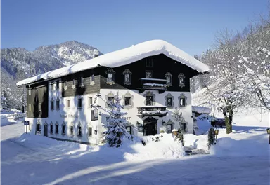 A traditional old house in the snow with a white roof. The surroundings are wintry and picturesque, surrounded by mountains and trees.