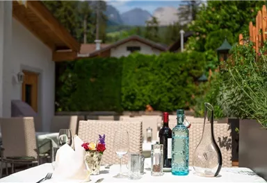 Ein schöner Tisch im Freien mit Gläsern, Flaschen und einer Blumendekoration. Im Hintergrund sind Bäume und Berge zu sehen.