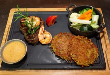 Ein köstliches Gericht mit Rinderfilet, Garnelen und knusprigen Kartoffelpuffern. Dazu gibt es frisches Gemüse und eine cremige Sauce.