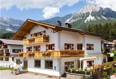 Ein charmantes Alpenhaus mit Holzbalkonen und schönen Fenstern. Im Hintergrund sind beeindruckende Berge und ein blauer Himmel zu sehen.