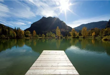 Ein ruhiger See mit einer Holzplattform im Vordergrund. Im Hintergrund sind Berge und bunte Herbstbäume zu sehen.