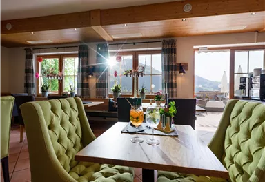 Ein gemütliches Restaurant mit grünen Stühlen und einem Holztisch. Auf dem Tisch stehen zwei Getränke und die Aussicht zeigt eine schöne Landschaft durch die Fenster.