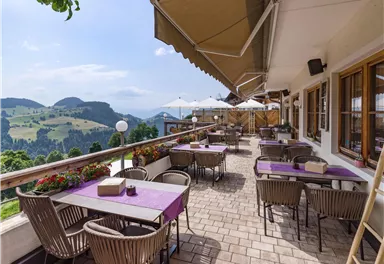 Eine einladende Terrasse mit Tischen und Stühlen, umgeben von blühenden Blumen. Im Hintergrund erstrecken sich sanfte Hügel unter einem klaren Himmel.