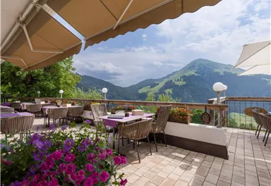 Eine schöne Terrasse mit Tischen und Stühlen, umgeben von bunten Blumen. Im Hintergrund sind grüne Berge und ein bewölkter Himmel zu sehen.