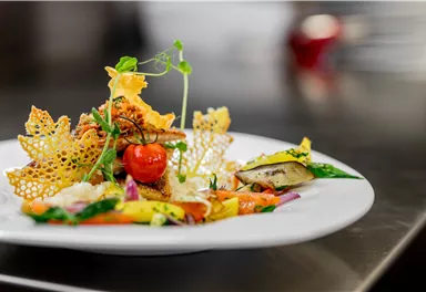 Ein kunstvoll angerichteter Teller mit gebratenem Gemüse, Fisch und einer knusprigen Dekoration. Die Farben sind lebhaft und ansprechend, was das Gericht sehr appetitlich wirken lässt.
