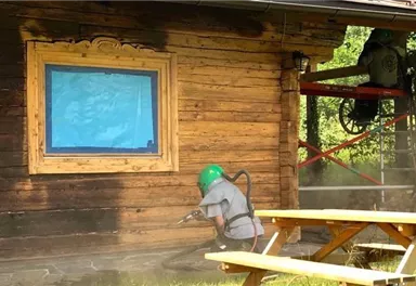 A craftsman is cleaning a wooden wall with a high-pressure device. In the background, a scaffolding and a blue window frame can be seen.