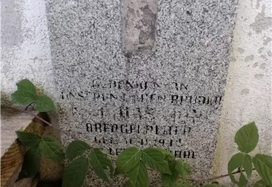 Eine Gedenktafel aus Grabsteinmaterial, teilweise von Pflanzen überwuchert. Der Text ist schwer lesbar und befindet sich an einer Wand.
