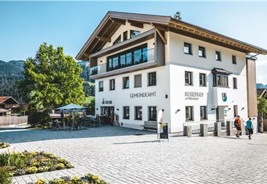 Ein modernes Gebäude mit der Aufschrift "Gemeindeamt". Vor dem Gebäude sind Bäume, Blumenbeete und einige Menschen zu sehen.