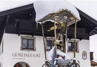 A fountain with snow on the roof and a statue in the foreground. In the background stands a traditional building with the inscription "Town Hall".