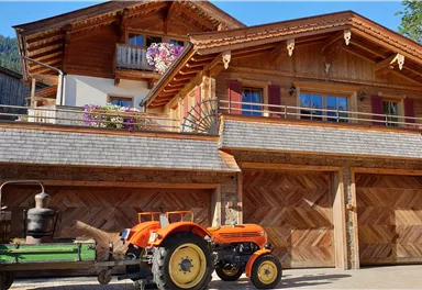 Ein rustikales Holzhaus mit schöner Architektur und Balkonen. Vor dem Haus steht ein orangefarbener Traktor auf einem gepflasterten Vorplatz.